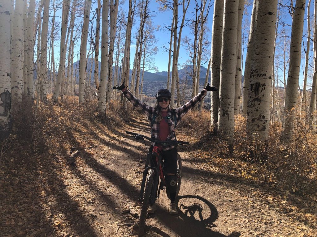 Thea, a young woman stands over a full suspension mountain bike, surrounded by Aspen trees in Moab, Utah. Her arms are raised in the air and excitement and she's enjoying a nice sunny beautiful day